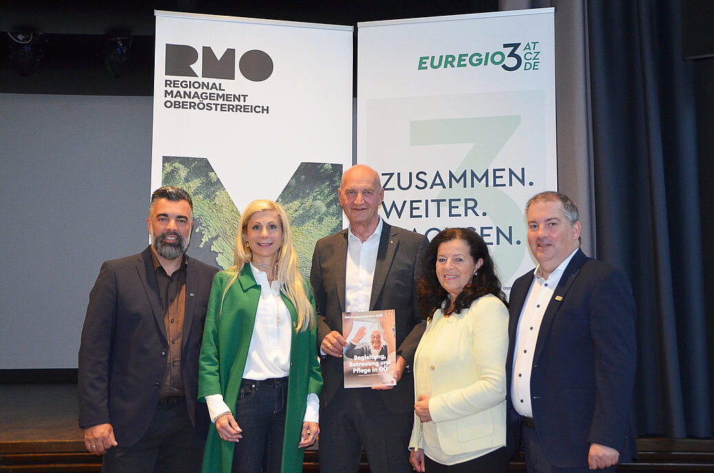 Fünf Personen stehen vor Roll‑ups von „RMO Regional Management Oberösterreich“ und „Euregio“. Eine Person hält eine Broschüre „Begleitung, Betreuung und Pflege in OÖ“ in der Hand.